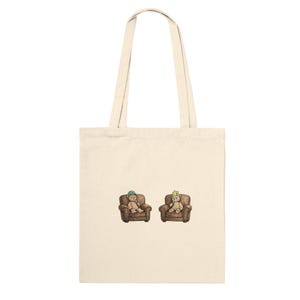 May include: Cream-colored tote bag with two teddy bears sitting in brown armchairs. One bear wears a green hat, the other a gold crown. The bag has long, cream-colored handles.