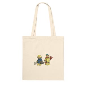 May include: Cream-colored tote bag featuring an illustration of two teddy bears dressed in rain gear. One bear holds a fishing net, the other a crab. The bears wear yellow boots and hats, and blue and yellow raincoats.