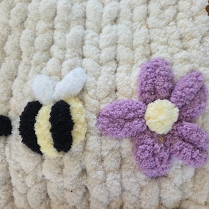 May include: A handmade, off-white, chunky knit blanket with a bee and flower design. The bee has black and yellow stripes with white wings. The flower is purple with a yellow center. The blanket is made of soft, textured yarn.