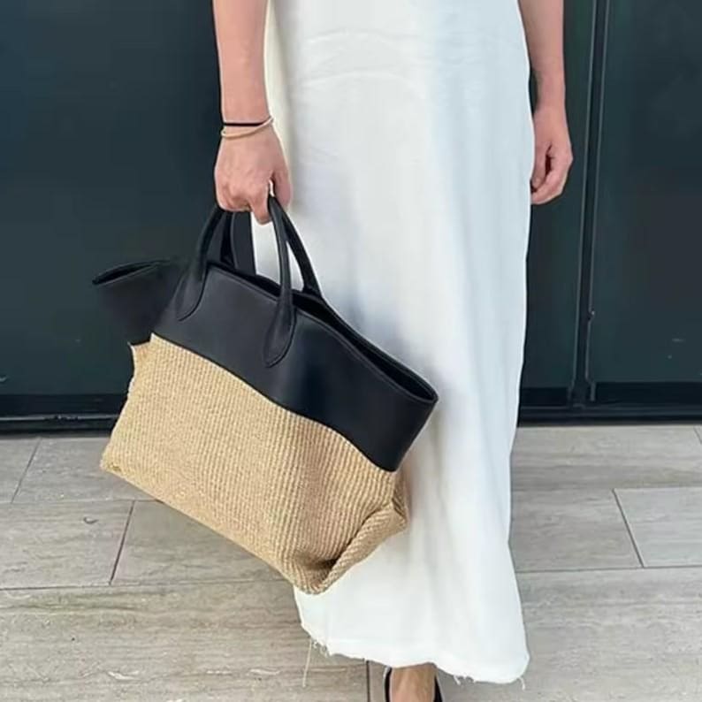 May include: A two-tone tote bag with a black leather top and handles, and a woven beige bottom. The bag is held by a person wearing a white dress.