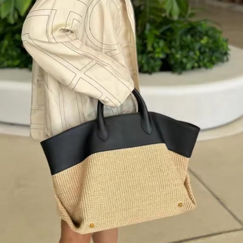May include: A large tote bag with a woven straw bottom and a black leather top. The bag has two black handles and gold-colored rivets at the bottom. The bag is being held by a person wearing a cream-colored jacket.