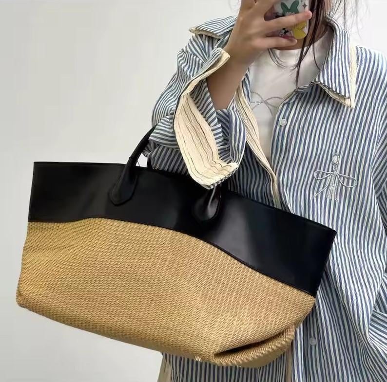 May include: A large tote bag with a woven straw bottom and a black leather top. The bag has two black handles and a spacious interior, perfect for carrying essentials. The bag's design is both stylish and functional.