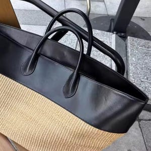 May include: Black and tan tote bag with two handles. The top half of the bag is black, and the bottom half is woven tan material. The bag has a zipper closure and is sitting on a surface.