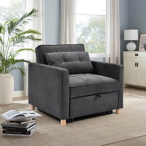 May include: A dark grey convertible armchair with a tufted seat and backrest, and a matching pillow. The chair has wooden legs and a pull-out bed feature. The room has a neutral colour palette with a large window and a potted plant.