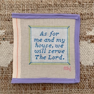 May include: A square, off-white needlepoint with the text "As for me and my house, we will serve The Lord." The text is in blue, within a green border, and framed by a lavender border. The background is a textured, tan surface.