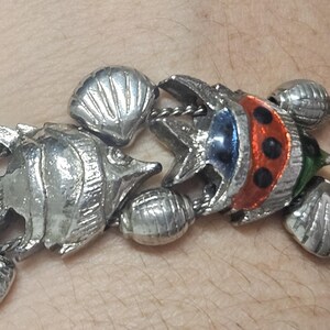 May include: Silver-toned bracelet featuring fish and seashell charms. The fish charms have red, blue, and green accents. The bracelet is connected with a chain and has a clasp closure. A nautical-themed jewelry piece.