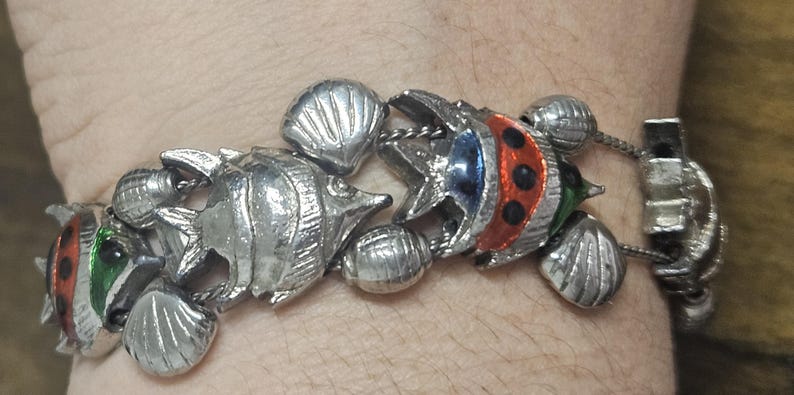 May include: Silver-toned bracelet featuring fish and seashell charms. The fish charms have red, blue, and green accents. The bracelet is connected with a chain and has a clasp closure. A nautical-themed jewelry piece.