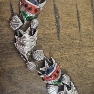 May include: Silver-toned necklace featuring a repeating pattern of stylized fish and seashells. The fish have red, black, green, and blue enamel details. The necklace is made of metal and has a delicate chain.