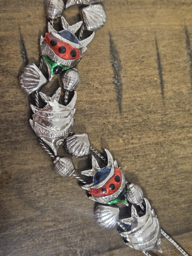 May include: Silver-toned necklace featuring a repeating pattern of stylized fish and seashells. The fish have red, black, green, and blue enamel details. The necklace is made of metal and has a delicate chain.