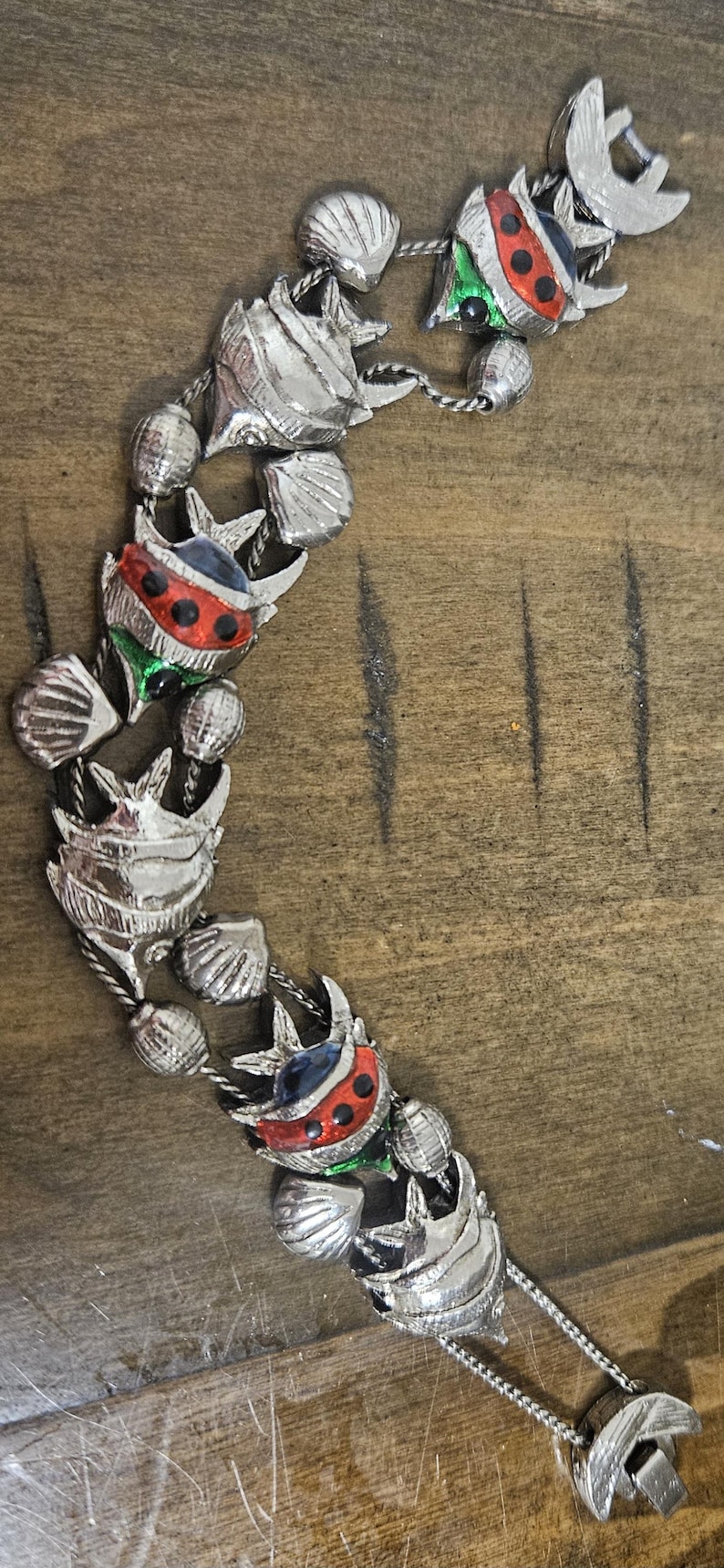 May include: Silver-toned bracelet featuring alternating seashell and ladybug designs. The ladybugs have red, green, and blue accents with black spots. The bracelet is on a wooden surface.