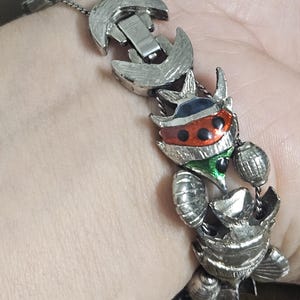 May include: Silver-toned metal bracelet featuring a series of stylized insect-like figures. The bracelet has a clasp closure and is accented with red, black, green, and blue enamel details. The bracelet is worn on a wrist.