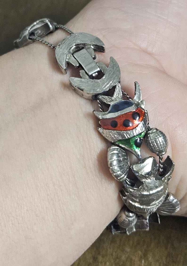 May include: Silver-toned metal bracelet featuring a series of stylized insect-like figures. The bracelet has a clasp closure and is accented with red, black, green, and blue enamel details. The bracelet is worn on a wrist.