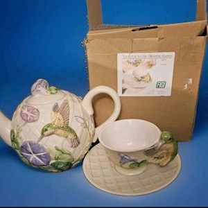 May include: A ceramic tea set with a teapot, teacup, and saucer. The set is decorated with hummingbirds, purple flowers, and green leaves. The teapot has a lid with a flower-shaped knob. The teacup has a hummingbird handle. The set is in a box.