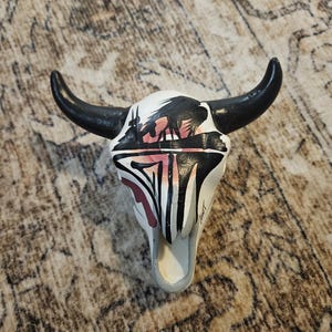 May include: A decorative white bull skull with black horns. The skull is painted with a black and pink design featuring a landscape scene and vertical stripes. The horns are curved and painted black.