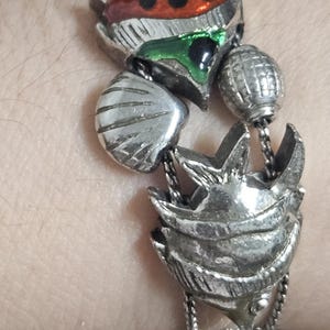 May include: Silver-toned metal bracelet featuring ornate, detailed charms. The bracelet includes a central charm with red, green, and blue accents, along with other decorative elements. The bracelet is designed with a unique, artistic style.