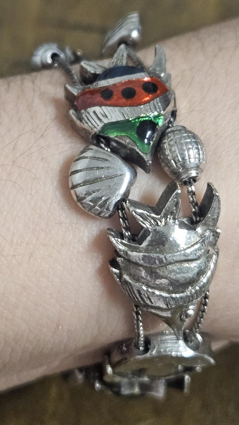 May include: Silver-toned metal bracelet featuring ornate, detailed charms. The bracelet includes a central charm with red, green, and blue accents, along with other decorative elements. The bracelet is designed with a unique, artistic style.