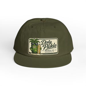 May include: An olive green baseball cap featuring a rectangular graphic on the front. The graphic depicts a smiling pickle wearing sunglasses and holding a surfboard, with the text Dirty Pickle BEACH CO. SARASOTA, FL printed next to it.