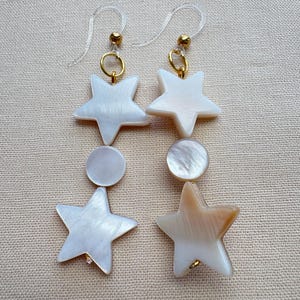 May include: A pair of drop earrings featuring iridescent white mother-of-pearl components. Each earring consists of a top star, a central circular disc, and a bottom star, all connected by gold-toned metal rings and clear plastic ear hooks.