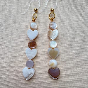 May include: A pair of long drop earrings featuring a vertical arrangement of alternating heart-shaped and circular shell beads in shades of white, cream, and brown. The earrings are attached to clear plastic ear hooks with small gold-tone metal rings.