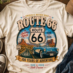 以下が含まれることがあります： クリーム色のTシャツ。ルート66のグラフィックが特徴で、青いクラシックカー、砂漠の風景、テキストが描かれています。「America's Mother Road」と「100 Years of Adventure」の文字が含まれています。茶色の野球帽と青いジーンズも表示されています。