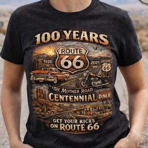 May include: A charcoal gray t-shirt featuring a graphic print celebrating 100 years of Route 66. The design includes a vintage car, a motorcycle, a desert landscape, and text reading 100 YEARS, ROUTE 66, THE MOTHER ROAD, CENTENNIAL DINER, and GET YOUR KICKS ON ROUTE 66.