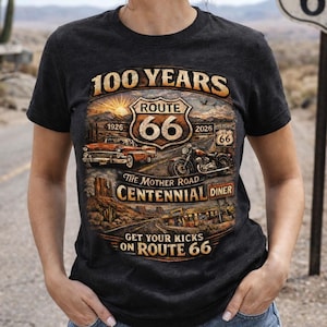 Puede incluir: Camiseta gris oscuro con un diseño de la Ruta 66 que incluye un coche clásico, una motocicleta y un restaurante. La camiseta conmemora los 100 años de la Ruta 66, con texto como "The Mother Road" y "Get Your Kicks on Route 66".