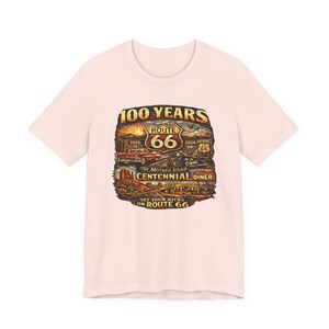 May include: A light pink t-shirt featuring a graphic print of a desert landscape with a red car, a motorcycle, and a Route 66 shield. The text reads 100 YEARS, ROUTE 66, 1926, 2026, THE MOTHER ROAD, CENTENNIAL DINER, and GET YOUR KICKS ON ROUTE 66.