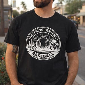 May include: Black t-shirt with a white graphic featuring a baseball, cactus, and palm trees, with the text "AZ SPRING TRAINING BASEBALL" in a circular design. The shirt is worn by a person wearing a tan baseball cap.