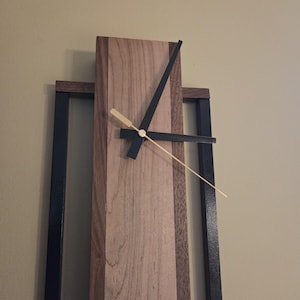 May include: A modern wooden clock with a light brown face and black hands. The clock's face is made of light and dark wood, and it is set in a black metal frame. The clock's hands are black and gold.