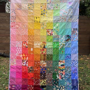 Op de afbeelding: Een regenboogdeken gemaakt van kleurrijke vierkante stoffen. Elk vierkant heeft een ander patroon, waaronder bloemen, geometrische en abstracte ontwerpen. Het deken is afgewerkt met een witte en blauwe bies.