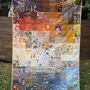 Könnte beinhalten: Eine große Patchwork-Decke mit einer Vielzahl von bunten Stoffen und Mustern. Die Decke besteht aus Quadraten in verschiedenen Größen und Farben, und die Muster umfassen geometrische Formen, Blumenmuster und abstrakte Kunst. Die Decke hängt an einem Holzzaun.
