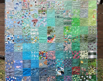 May include: A large quilt made of many different fabric squares. The squares are arranged in a gradient pattern, starting with light green and transitioning to dark blue. The quilt is made of a variety of fabrics, including floral prints, geometric patterns, and solid colors.