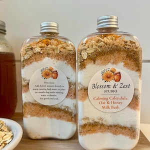 May include: Two clear plastic bottles filled with layered bath soak ingredients including oats, dried calendula flowers, and white powder. The labels read Blossom & Zest Studio Calming Calendula Oat & Honey Milk Bath with usage directions.