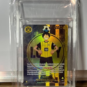 Peut inclure: Une carte à collectionner dans un étui protecteur, représentant un personnage en maillot de foot jaune et noir. La carte affiche le texte "LUCY", "BVB 09" et "1&1". Un ballon de foot est au-dessus de la tête du personnage.
