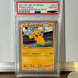 Könnte beinhalten: Eine bewertete Pokémon-Karte mit Pikachu in einer Fußball-Illustration. Die Karte ist mit "2014 P.M. JPN. XY PROMO PIKACHU-HOLO J-LEAGUE SOCCER-1st PRINT" beschriftet und mit GEM MT 10 bewertet. Die Karte hat japanischen Text.