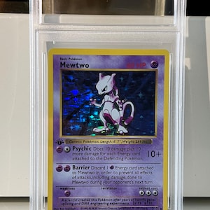 Può includere: Una carta Pokémon Mewtwo valutata in una custodia protettiva. La carta presenta un'immagine olografica di Mewtwo, una creatura viola e bianca. Il testo include "Basic Pokémon Mewtwo" e dettagli del gioco. La carta è lunga 201 cm e pesa 122 kg.