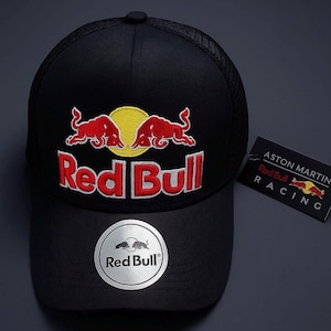 Op de afbeelding: Zwarte baseballcap met het Red Bull-logo geborduurd in rood en geel. De cap heeft een mesh achterkant en een gebogen klep. Een zilveren sticker met het Red Bull-logo staat op de voorkant. Een label met het Aston Martin Red Bull Racing-logo is bevestigd.