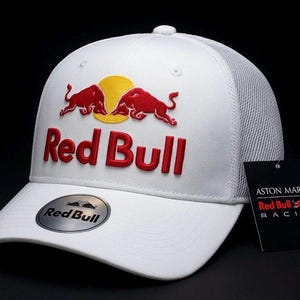 Könnte beinhalten: Weiße Baseballkappe mit dem Red Bull-Logo in Rot und Gelb. Die Kappe hat einen Mesh-Rücken und einen gebogenen Schirm. Ein kleines Metallabzeichen und ein schwarzes Etikett mit dem Aston Martin Red Bull Racing-Logo sind ebenfalls sichtbar.