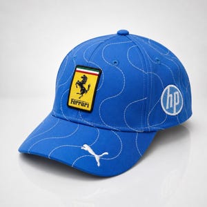 Può includere: Un cappellino da baseball blu con cuciture bianche e visiera curva. Il cappellino presenta una toppa con il logo Ferrari, un logo Puma e un logo HP. Il cappellino è realizzato in tessuto resistente e ha un bottone sulla parte superiore.