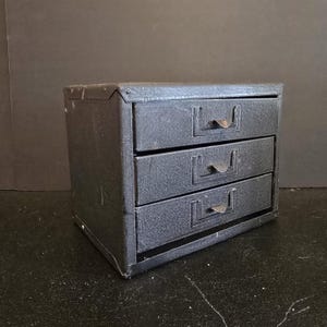 May include: A small, vintage, three-drawer cabinet in a dark gray color. The cabinet is made of metal with riveted corners and small, curved metal drawer pulls. The drawers are rectangular and have a textured surface.