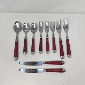 May include: A set of flatware consisting of three spoons, four forks, and two knives. Each piece features a polished silver-tone metal head and a solid red handle secured with two small metal rivets. The utensils are arranged in a neat formation.