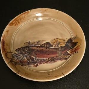 May include: A beige ceramic plate featuring a detailed illustration of a brown trout. The fish is depicted with reddish-brown and black markings, surrounded by aquatic plants and rocks. The plate has a brown rim and the artist's signature.
