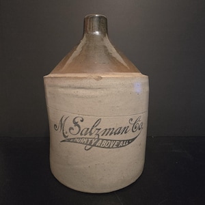 May include: A vintage stoneware jug with a rounded body and a narrow neck. The jug is primarily beige with a brown top and a dark brown spout. The words "M. Salzman Co." and "Purity Above All" are printed in black script on the front.