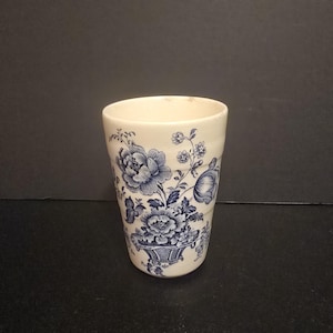 May include: Cream-colored ceramic tumbler with a blue floral design. The design features large peony-like flowers and smaller blossoms arranged in a basket. The tumbler has a slightly flared shape and a smooth, glossy finish.