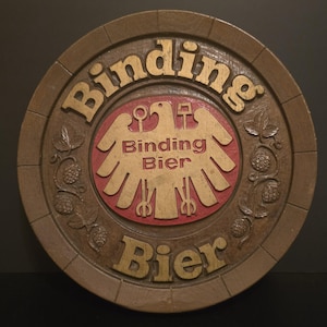 May include: A round, wooden sign with the words "Binding Bier" in gold lettering. The center features a red and gold emblem with an eagle and the words "Binding Bier". Hops and leaves are carved around the edge.