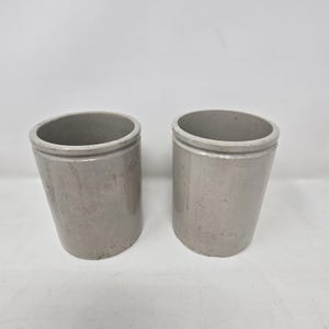 May include: Two cylindrical ceramic crocks with a smooth, light gray glazed finish. Each vessel features a simple, open-top design with a slightly flared rim and a solid, uniform structure, presenting a neutral appearance suitable for various storage.