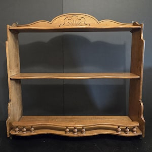 May include: A light brown wooden wall shelf with three tiers. The top shelf has a decorative, scalloped edge with carved details. The bottom shelf features small, rounded pegs. The shelf is designed for displaying items.