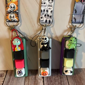 May include: Three lanyards with keychains featuring characters from The Nightmare Before Christmas. The lanyards have different patterns, including Jack Skellington faces and other characters. The keychains have colourful, stacked keycaps with character designs.