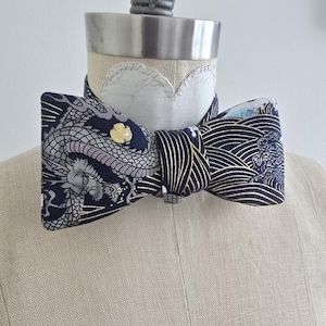 May include: A navy blue bow tie with a detailed dragon and wave pattern. The bow tie is displayed on a mannequin. The fabric features a dragon design in grey and pink, with a gold flower accent. The other side has a white and gold wave pattern.