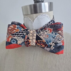 May include: A patterned bow tie with a floral and geometric design in shades of blue, red, brown, and beige. The bow tie is displayed on a mannequin head, showcasing its shape and style. The fabric has a patchwork appearance.